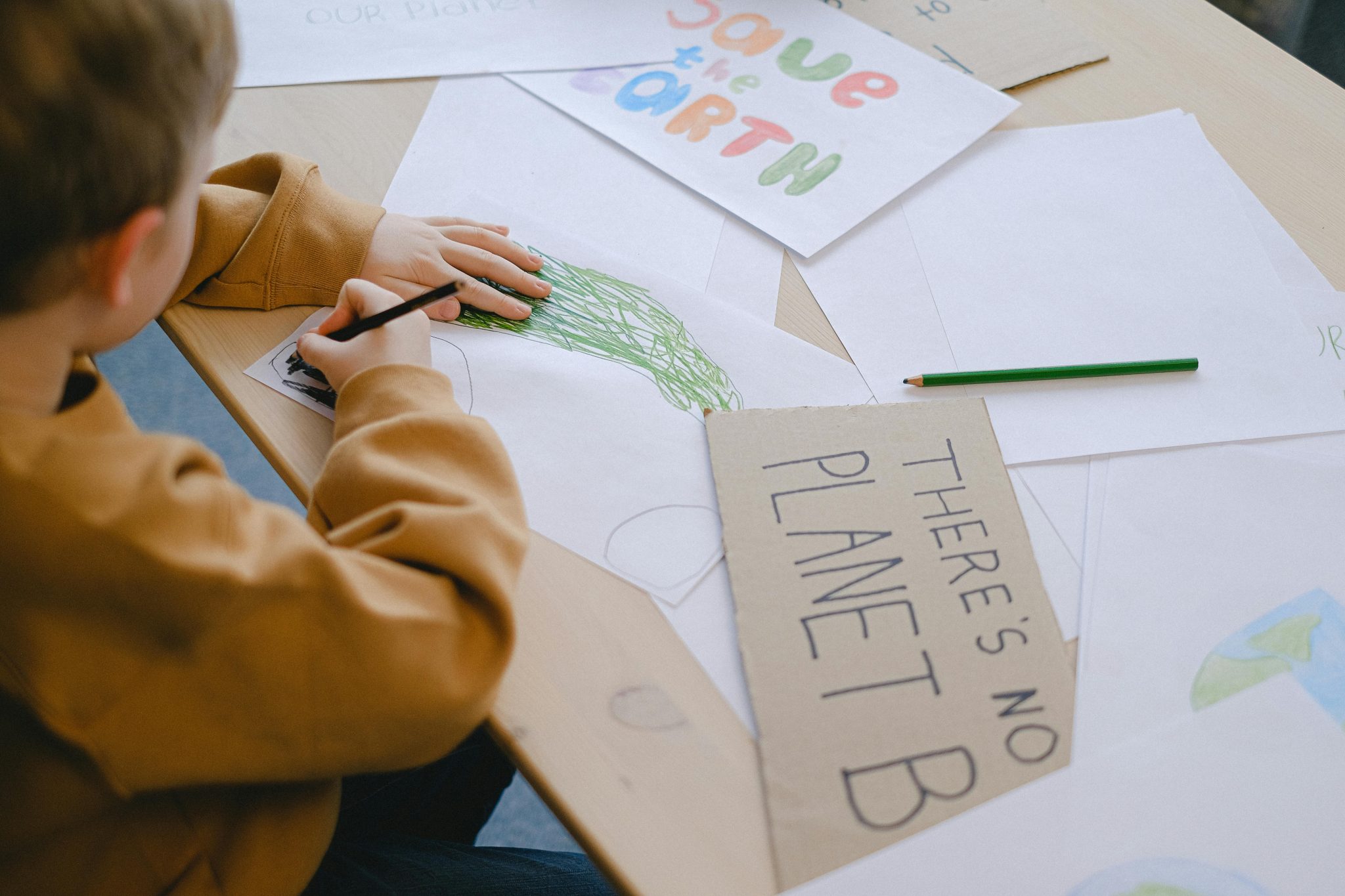 Băiat care desenează la birou mesaje sustenabile și îndemnuri pentru protejarea planetei. Sursa foto:Anna Shvets/Pexels